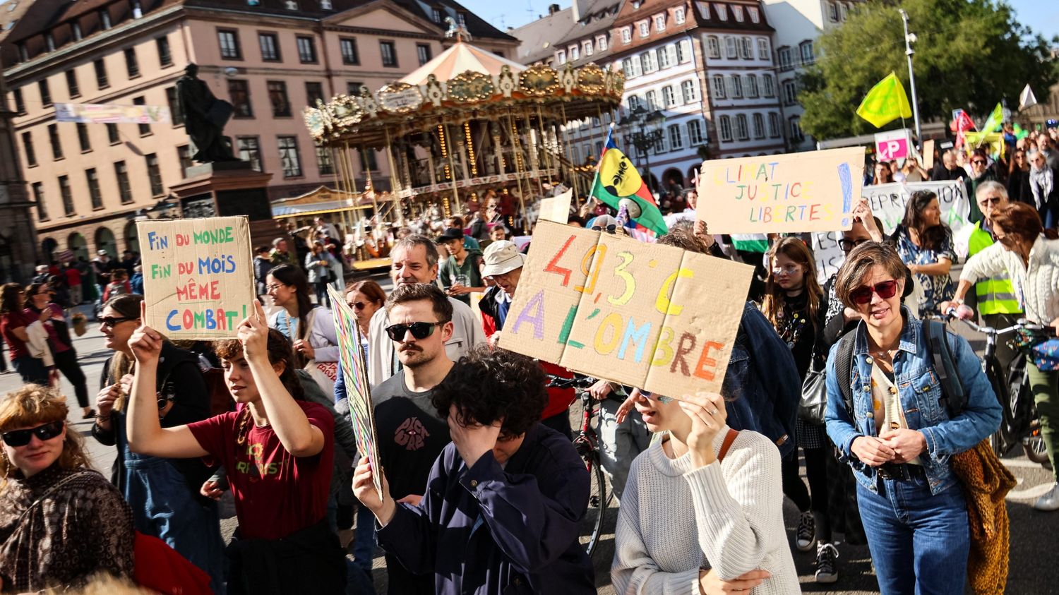 Une marche pour le climat place Broglie à Strasbourg : dans la foule, une jeune femme brandit une pancarte en carton « Fin du monde, fin du mois, même combat ! », une autre « 49,3 °C à l'ombre », une troisième « Climat, justice, libertés ! ». Derrière, un manège et les façades de la place.