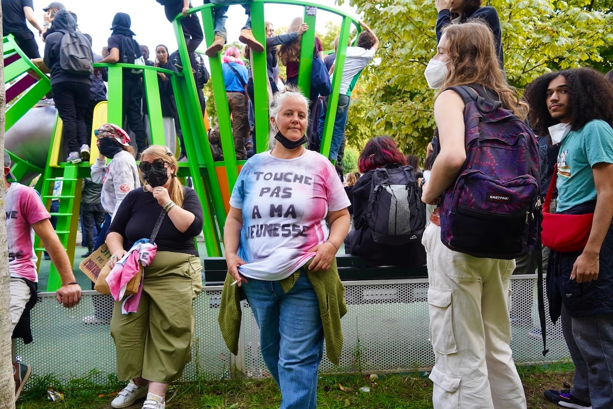 Une manifestation de lycéen·nes à Paris : au premier plan, une femme porte un t-shirt peint « Touche pas à ma jeunesse » ; derrière elle, des jeunes grimpés sur une aire de jeux.