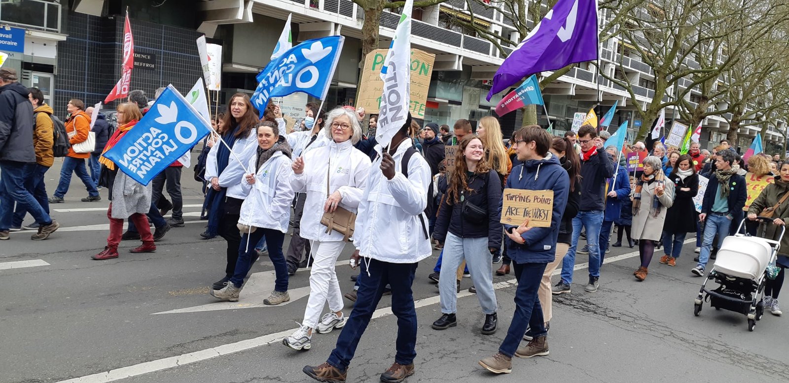 Des bénévoles d'Action contre la Faim en veste blanche défilent dans une marche pour le climat, drapeaux bleus de l'association levés. Autour d'eux, la foule, une pancarte en carton « Tipping points are the worst », des drapeaux violets et une poussette.