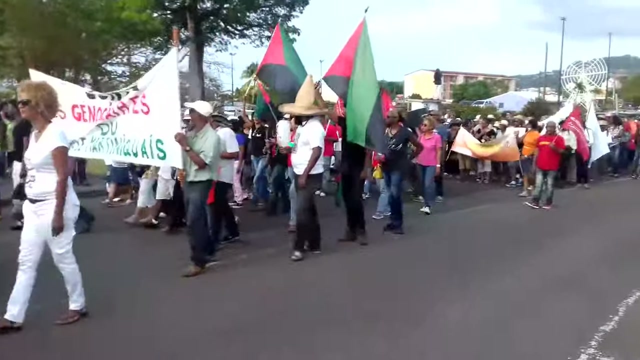 Un cortège dans une rue du Lamentin, en Martinique. Au premier plan, une banderole blanche dénonce les « génocidaires du peuple martiniquais » ; des drapeaux rouge, vert et noir flottent au-dessus des manifestantes et manifestants.