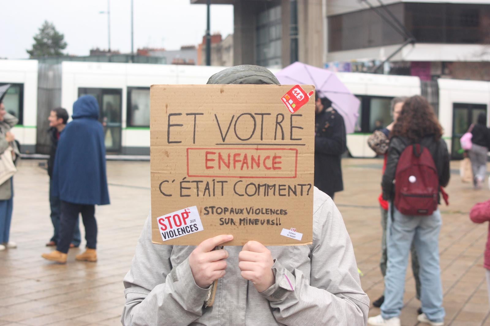 Une personne cache son visage derrière une pancarte en carton : « Et votre enfance, c'était comment ? Stop aux violences sur mineurs !! », avec des autocollants du 119.
